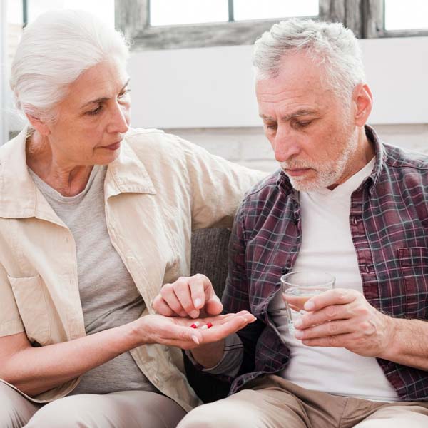 An older woman holds an older man's pills as he takes them one by one with the water he's holding.