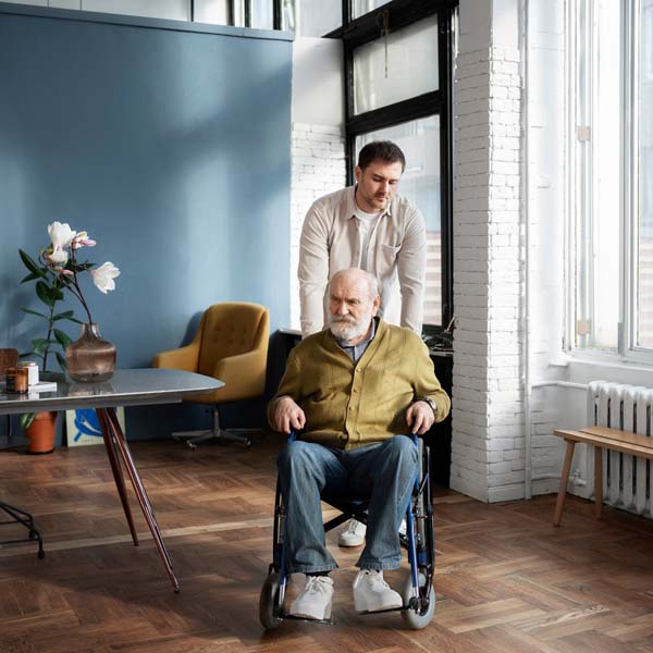 An older man gets pushed on his wheel chair in his residence by a younger man.