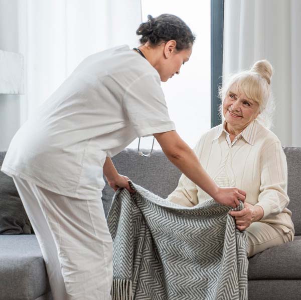 A caregiver handing a blanket to an older woman sitting on a couch.