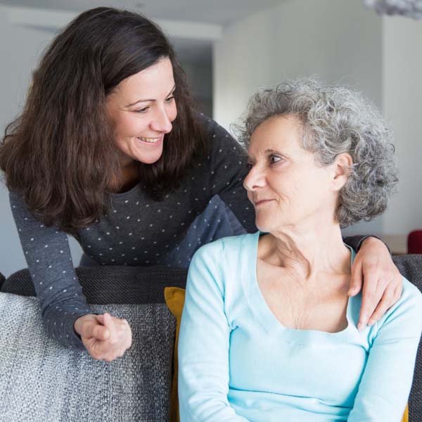 An older woman in her residence smiles at a younger woman who has her hand placed on the older woman's shoulder while smiling.
