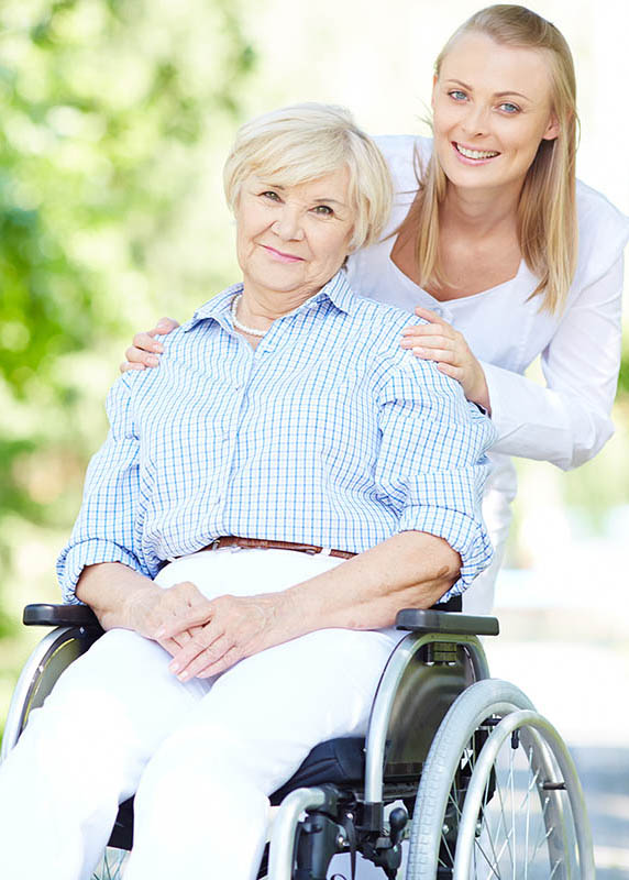 An older woman sitting in a wheel chair and a younger woman behind her with her hands placed on the older woman's shoulders. Both are smiling.